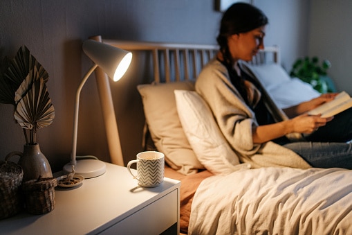 Hot tea in a cup on a night stand. On the bed a woman is reading a book in cozy clothes.